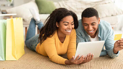 Cheerful millennial african american couple with bags with purchases lying on floor with tablet,...
