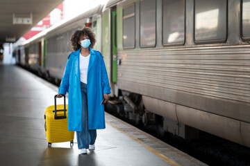 Young Black Woman Wearing Medical Mask Walking With Suitcase At Railway Station
