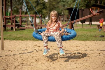 Happy cheerful trendy elementary school age girl, child sitting on a modern swing outdoors, outside, one person, real people lifestyle. Playground fun, cool kid resting on a swing, taking a break