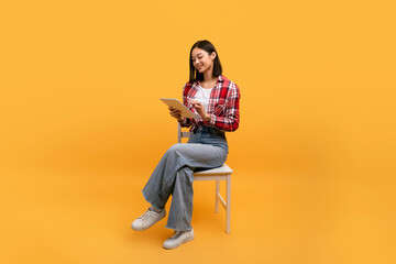 Young asian lady using digital tablet, sitting on chair, enjoying modern technologies, yellow background