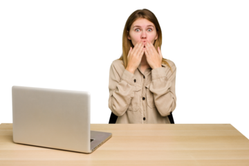 Young caucasian woman in a workplace working with a laptop isolated shocked, covering mouth with hands, anxious to discover something new.