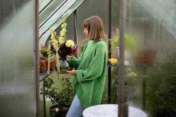 Young woman putting bouquet of flowers on shelf in tiny orangery at backyard. Vintage greenhouse made of rusty metal and glass