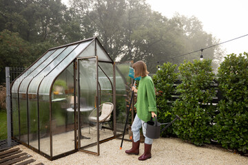 Woman with rag and watering can near greenhouse for growing plants at backyard. Gardening, leisure time in garden concept