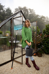 Portrait of young woman stands with rag and watering can near greenhouse for growing plants at backyard. Gardening, leisure time in garden concept