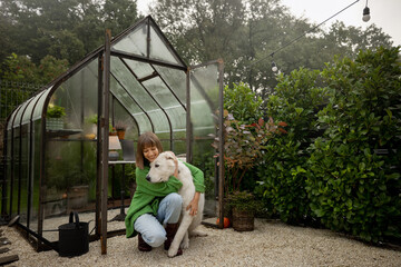 Young woman with her white dog near glasshouse at backyard, spending leisure time together outdoors