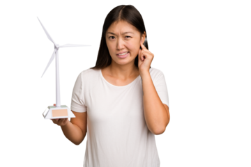 Young asian woman holding a small wind energy mill isolated covering ears with hands.