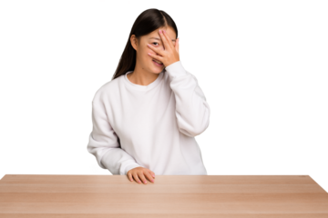 Young asian woman sitting on a table isolated blink at the camera through fingers, embarrassed covering face.
