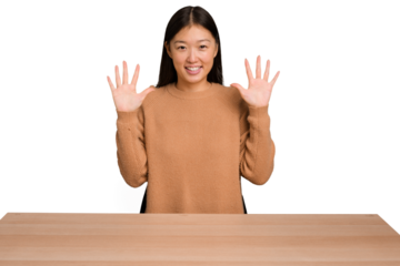 Young asian woman sitting on a table isolated showing number ten with hands.