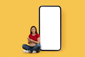 Mobile Mockup. Young Pregnant Woman With Smartphone Sitting Near Big Blank Telephone