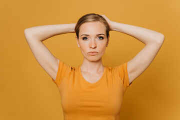Fototapeta premium Beautiful young red haired woman in orange t-shirt, collects her hair on the head by both hands, looks confident, over yellow background. Purposeful people concept.