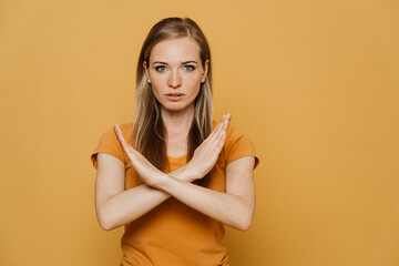 Obraz premium Confident redhead pretty woman in orange t-shirt, shows gesture sign stop, crossing her arms, wants to stop the coronavirus pandemic. Stay home and health care concept. Stands over yellow background.