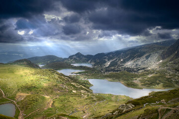 Dramatic dark blue sky over mountain glacial lakes in the Rila mountains, light leaks sunlight shining through clouds. 