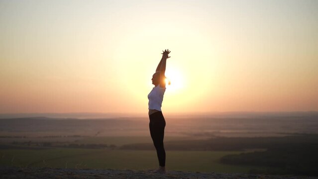 Silhouette of unrecognizable young meditate woman practicing yoga, bending forward and backward and finishing with namaste standing on top of mountains with beautiful summer landscape during sunset.