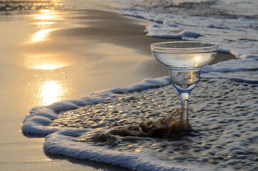 Martini in a glass goblet against the background of sand and waves on the seashore at sunrise.