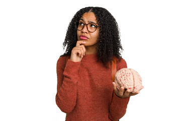 Young student african american woman holding a brain isolated looking sideways with doubtful and...