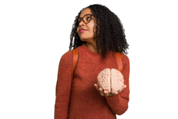 Young student african american woman holding a brain isolated looks aside smiling, cheerful and pleasant.