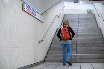 Rear view of single young woman with red backpack walking in the metro underground station with direction signs to diverse train on the wall
