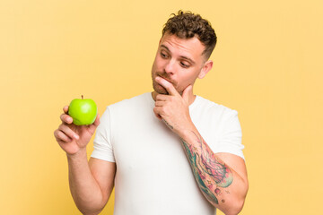 Young caucasian man eating an apple isolated looking sideways with doubtful and skeptical...