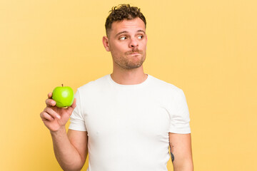 Young caucasian man eating an apple isolated confused, feels doubtful and unsure.