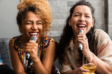 Girl friends sing karaoke excitedly in the living room - obrazy, fototapety, plakaty