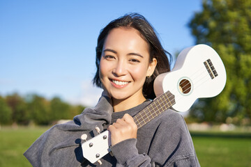 Portrait of beautiful smiling girl with ukulele, asian woman with musical instrument posing outdoors in green park - obrazy, fototapety, plakaty