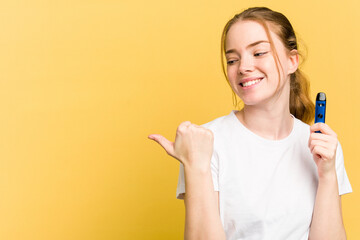 Young cute woman holding a vaper isolated on yellow background points with thumb finger away, laughing and carefree.