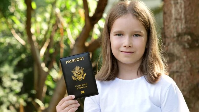 Happy cheerful kid, girl, young Eastern European, Caucasian child holding a US passport in hands, portrait, outdoors. Legal USA emigration, immigration and children simple concept, face closeup detail