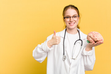 Young doctor caucasian woman holding a brain isolated on yellow background smiling and raising thumb up