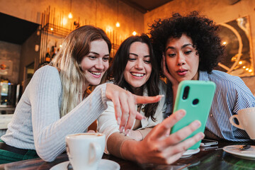 Group of young women having fun sharing media with an cellphone. Three girls looking to the smartphone on a coffee shop, restaurant or bar. Female students buying on a market place using a mobile - obrazy, fototapety, plakaty