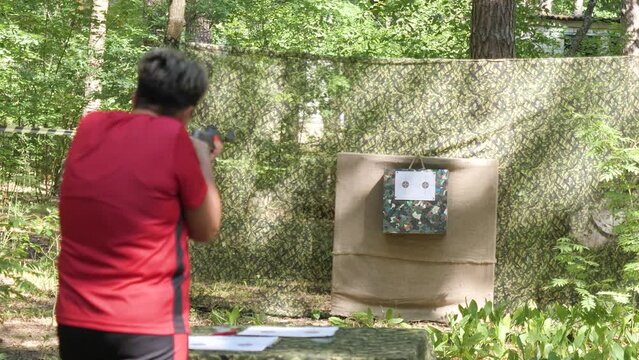 Closeup of woman with short hair in red t-shirt fires a rifle at shooting range target outside on summer day