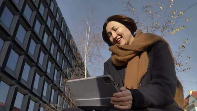 stylish fifty years old woman in scart and coat sits on bench with tablet in Wroclaw, Poland