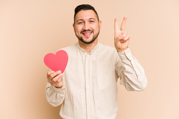 Adult latin man holding a red heart isolated showing number two with fingers.