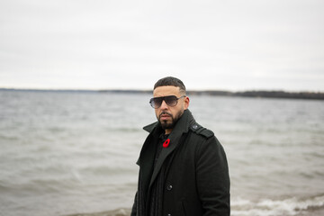 Bi racial model standing on board walk with water behind him winter time with sunglasses and green jacket on  - Powered by Adobe