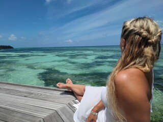 Blonde Frau im weißen Kleid sitzt auf Steg und schaut entspannt auf das weite türkisene Meer - Traumurlaub Malediven - Woman with blond hair is sitting on the turquoise beach and relaxing 