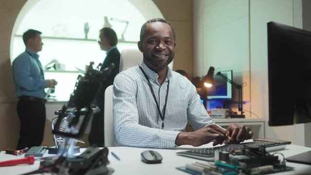 Portrait of young happy African robotics engineer working on computer coding programming in office. Attractive professional technician looking at camera smiling. Technologies concept.