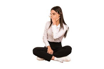 Young colombian woman sitting on the floor isolated blink at the camera through fingers,...