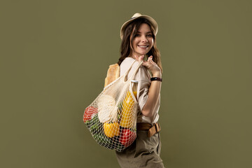 Smiling young woman in light summer clothes with a mesh eco bag full of vegetables, greens on a green studio background. Sustainable lifestyle. Eco friendly concept. Zero waste.