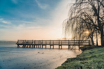 Jezioro zimą wczesnym rankiem z zimowymi mgłami w Zebrzydowicach na Śląsku w Polsce z parkiem pałacowym, pomost i zamarznięty basen.