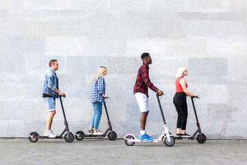 interracial company of friends rides electric scooters against the background of a wall, people use...