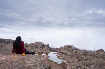 half-sitting Latin woman with her arms supporting her, with a lagoon and a valley of rocks in the background.