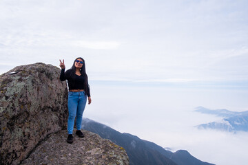 latin woman doing the peace and love pose with her hand, standing on the edge of a cliff with a cloud bed in the background.