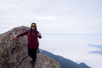 latin woman doing the metal hand pose, standing on the edge of a cliff with a cloud bed in the background.