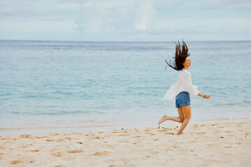 Naklejka premium Sports woman runs along the beach in summer clothes on the sand in a yellow T-shirt and denim shorts white shirt flying hair ocean view, beach vacation