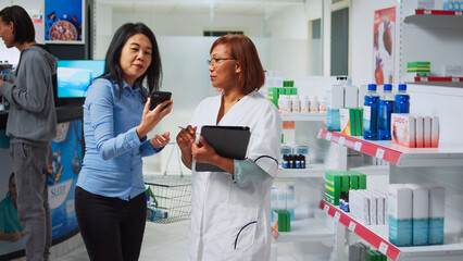 Medical consultant using digital gadget to do medicine inventory, analyzing pharmaceutical products on drugstore shelves. Woman working as pharmacist looking at pharmacy supplies stock.