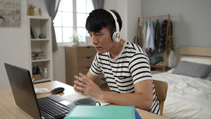 concentrated asian male student with a headset is writing notes and typing on the laptop while learning from home in the bedroom during lockdown