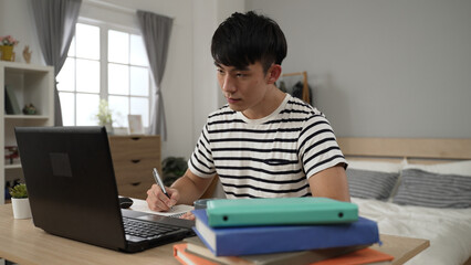 happy asian Korean college boy learning from home is typing on the laptop and writing in the...