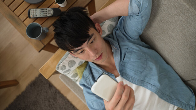 asian Japanese male is lying on comfortable couch scrolling on social media in the living room at home