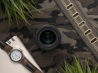 Overhead flat lay of a 30mm f1.4 camera lens surrounded by personal items on a camouflage background