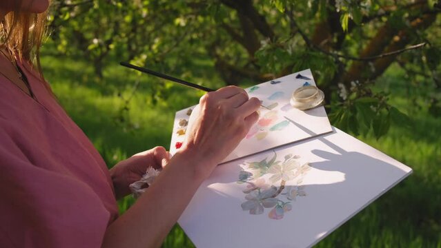 The artist paints in beautiful blooming gardens.Girl, an artist, paints bright, colorful flowers with delicate brush, watercolor or oil on canvas. Garden or park resort. Rest, relaxed mood, sunny day.