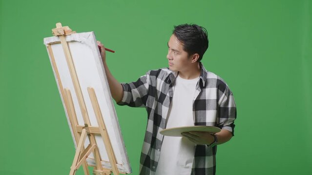 Asian Male Artist Is Concentrated Painting On Canvas By Oil Paints And Brush In The Green Screen Studio
